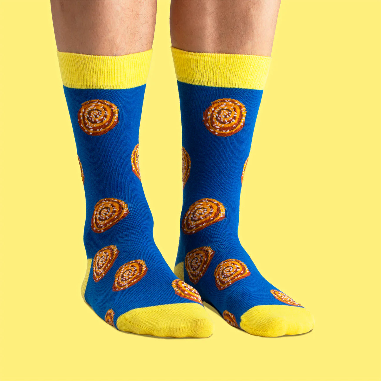 A pair of Svensk Husman Socks from the Fika Coffee Table Gift Box—blue with yellow accents and a cinnamon bun pattern—perfect for coffee lovers, pictured on a yellow background. Set includes 4 designs: Cinnamon Bun, Coffee Mug, Semla, and Biscuits.