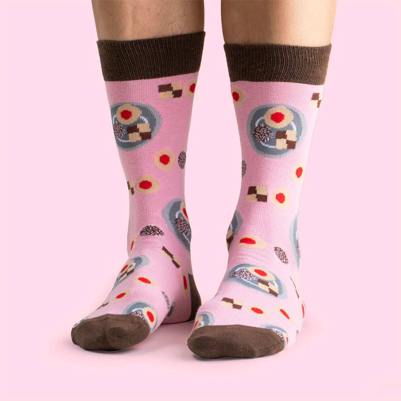 A person wears Svensk Husman Socks from the Fika Coffee Table Gift Box (set of 4, featuring cinnamon bun, coffee mug, semla, and biscuit patterns) with brown toes/heels/cuffs, standing on a light pink background—great for coffee lovers.