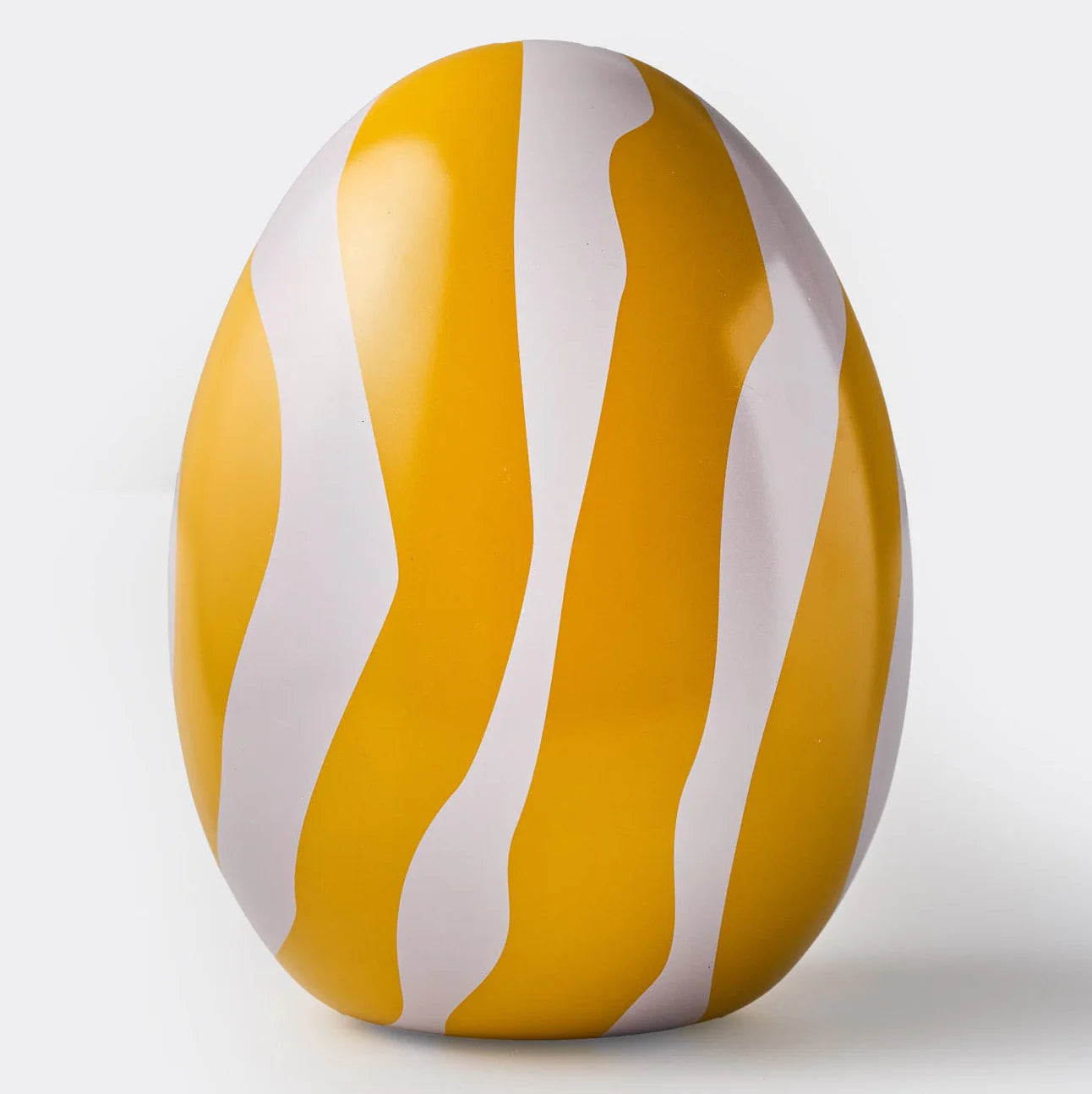A smooth, glossy Zebran Big Reusable Easter Egg by Gåva Gifting, decorated with bold yellow and white wavy stripes, stands upright against a plain light gray background, resembling a classic Swedish-style Påskägg in tin.