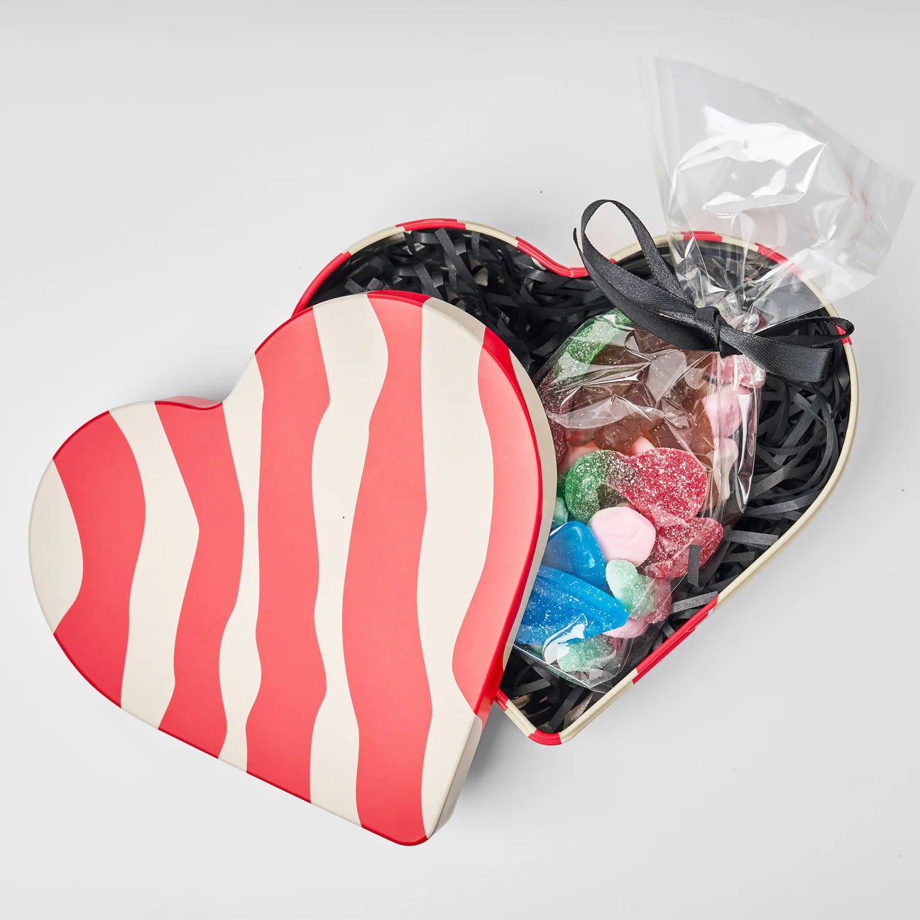 The Heart Reusable Tin - Älskar (Love) by Gåva Gifting features red and white wavy stripes, black shredded paper, and a clear bag of assorted Swedish candy tied with a black ribbon—perfect for sweet surprises.