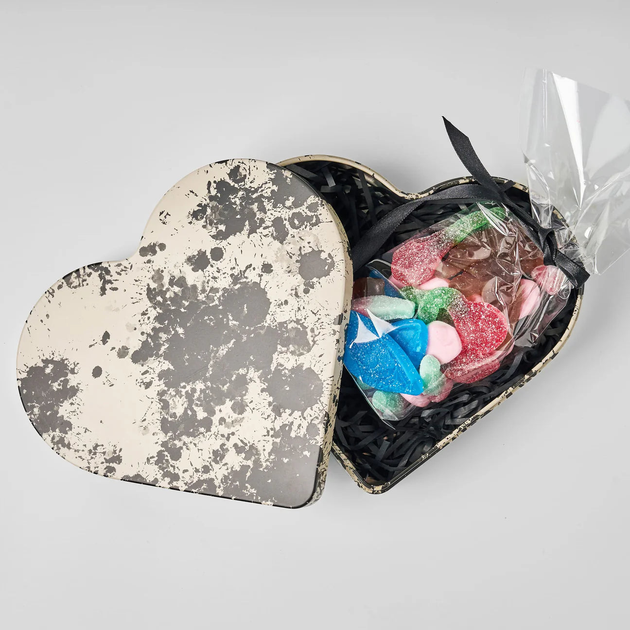 The Heart Reusable Tin - Tack (Thank You) by Gåva Gifting features a Swedish design heart-shaped tin with a black and white lid, paired with colorful heart gummy candies, all arranged on black shredded paper for a charming gift.