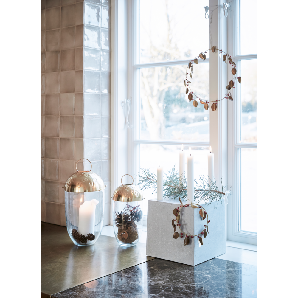 A decorative Christmas Wreath Leafi Auric in Gold, 12 CM by House Doctor hangs in a window above a sill featuring two glass lanterns with candles and a concrete block with a second handmade Christmas wreath and tall candles.