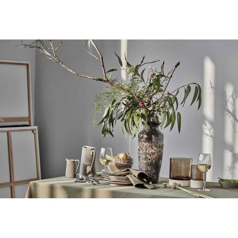 A dining table set with neutral-toned dishes, mouthblown glassware, and utensils features a large rustic vase with green branches. The handmade Candle Holder Rivo with brown glass from House Doctor adds a touch of elegance as sunlight casts shadows on the wall and table, creating a serene atmosphere.