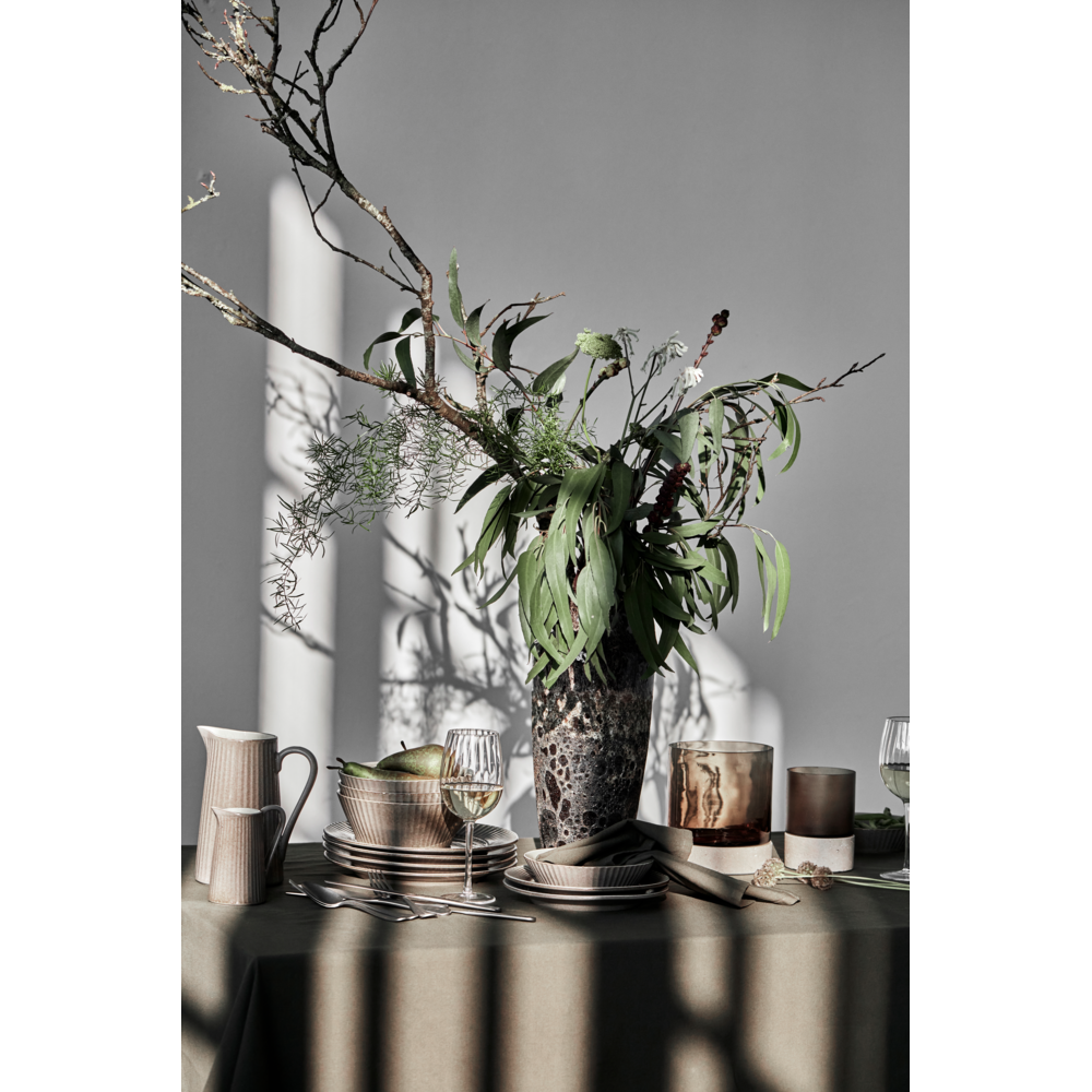 A table set with dishes, glasses, and a beige pitcher features a tall vase with green foliage and branches. The House Doctor Candle Holder Rivo with brown glass, handmade, sits elegantly beside mouthblown glassware as sunlight casts dramatic shadows on the table and wall behind.