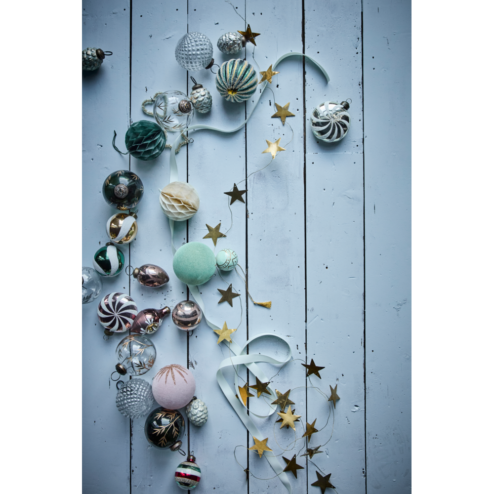A House Doctor Garland Stars in Antique Brass, 1.5 m, featuring handcrafted gold-colored stars interspersed with an assortment of spherical and patterned Christmas ornaments on a light blue wooden surface.