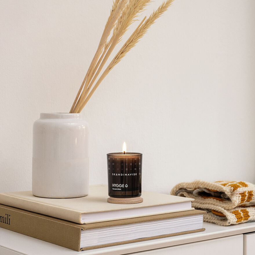 A Skandinavisk Mini Scented Candle Hygge 20h, 65g in a glass holder sits on stacked books beside a white vase with dried grasses and a folded knitted blanket, creating a cozy atmosphere on the white surface.