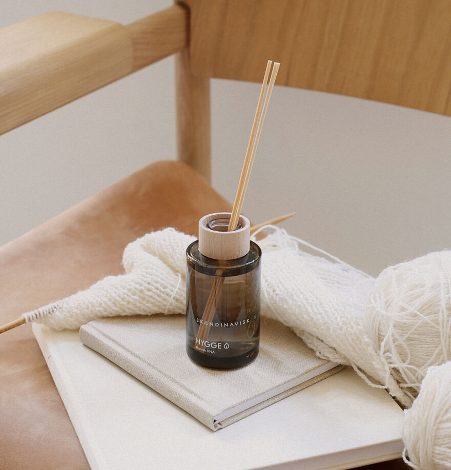 The Skandinavisk Scent Diffuser Hygge (Small) 100 ml sits on a closed book beside white yarn and needles, arranged on a tan chair, infusing the space with a cozy Scandinavian fragrance for Danish hygge.