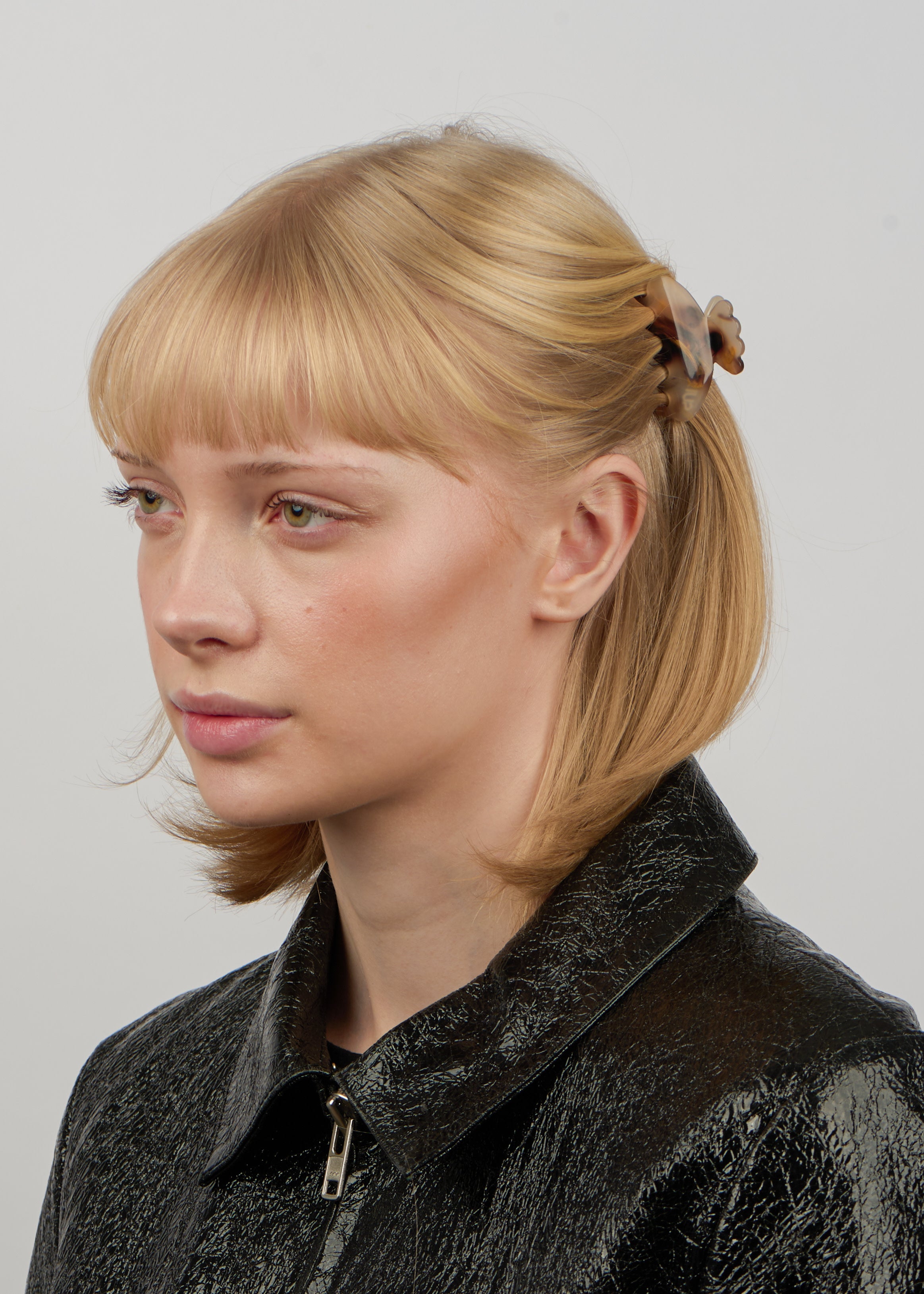 A woman with straight light brown hair, styled with the Sui Ava Helle Pretty Mini Hair Claw Clip in Champagne, wears a shiny black textured jacket and looks slightly to the left against a plain light background.