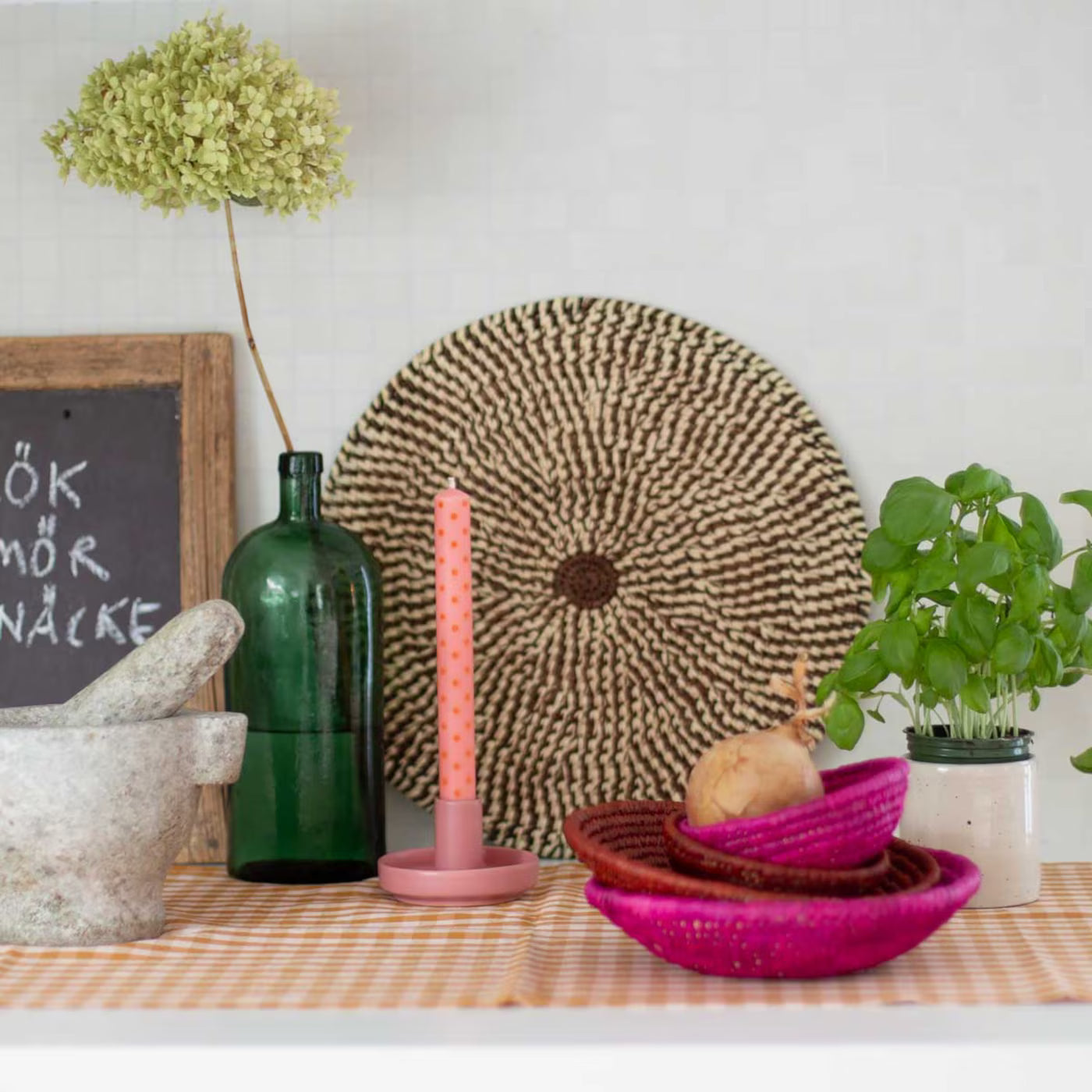 A kitchen shelf with a green bottle, a potted basil plant, stoneware mortar and pestle, woven baskets, dried flowers on gingham cloth, a framed chalkboard, and a pink Hermes handmade candle holder by A World of Craft.