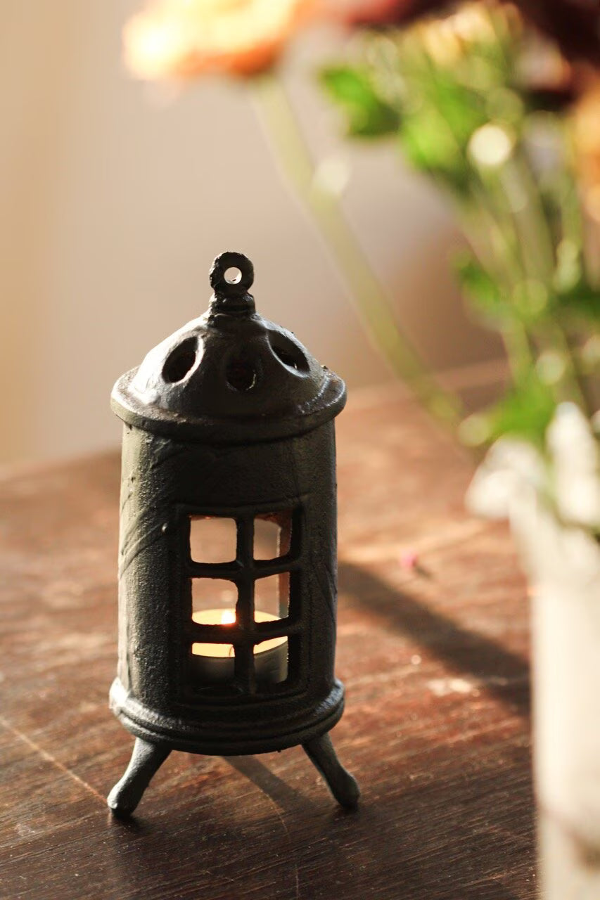 The Strömshaga Stove Candle Holder, crafted from cast iron, holds a lit candle on a wooden surface. Green leaves and orange flowers blur softly in the sunlit background, creating a warm, vintage-inspired home decor setting.