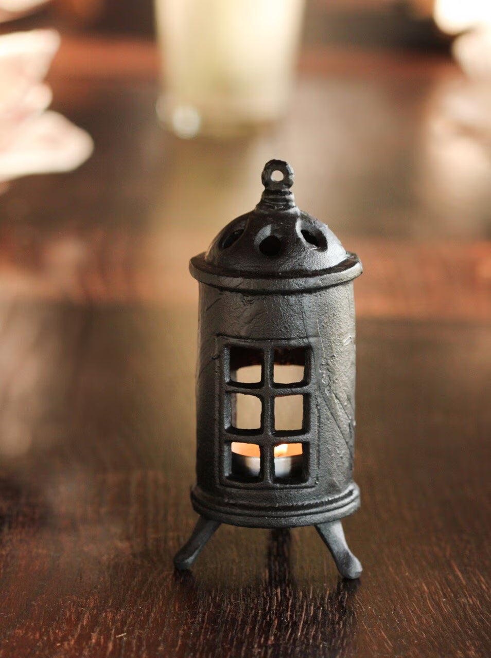The Strömshaga Stove Candle Holder, crafted from black cast iron with window cutouts, sits on a dark wood table. A candle inside gently illuminates this cozy decor piece, highlighting its charming lantern details against a blurred background.