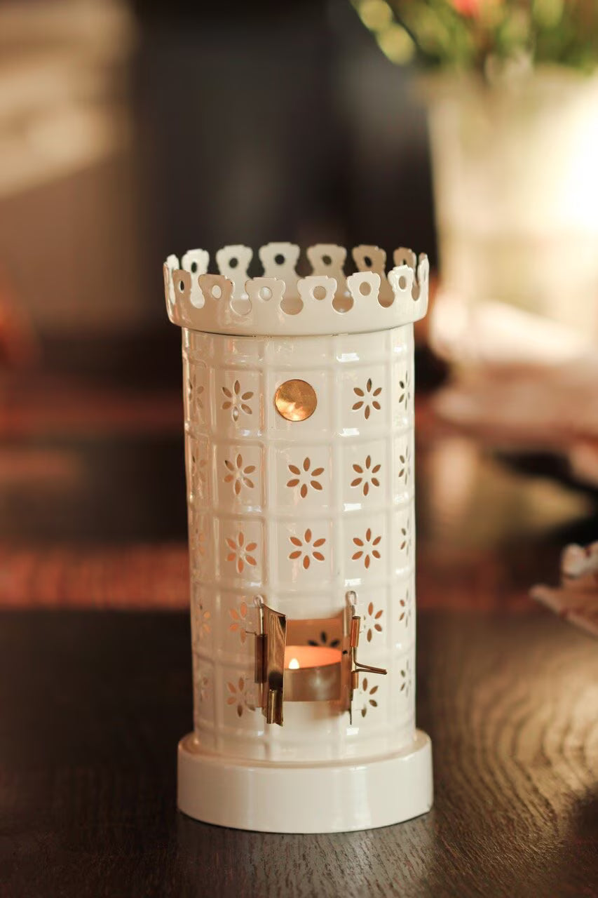 The Strömshaga Tea Light Holder, Tiled Stove in White, adds a decorative glow on a wooden surface with its charming ceramic design and flower-shaped cutouts, softly illuminating the blurred background.