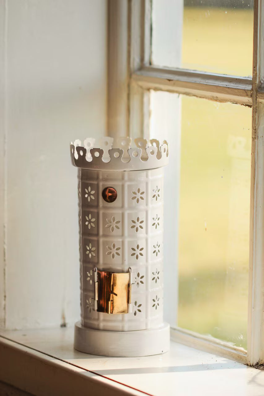 The Strömshaga Tea Light Holder, Tiled Stove in White, features a decorative design and gold accent, placed on a windowsill in soft natural light.