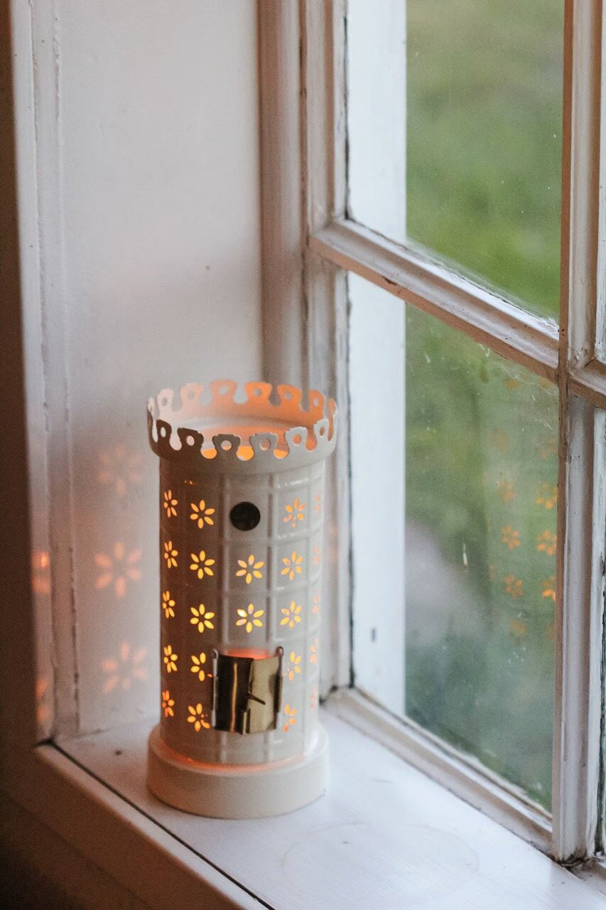 The Strömshaga Tea Light Holder, Tiled Stove in White, glows warmly on a windowsill with leafy views outside, its flower-shaped cutouts casting delicate patterns.