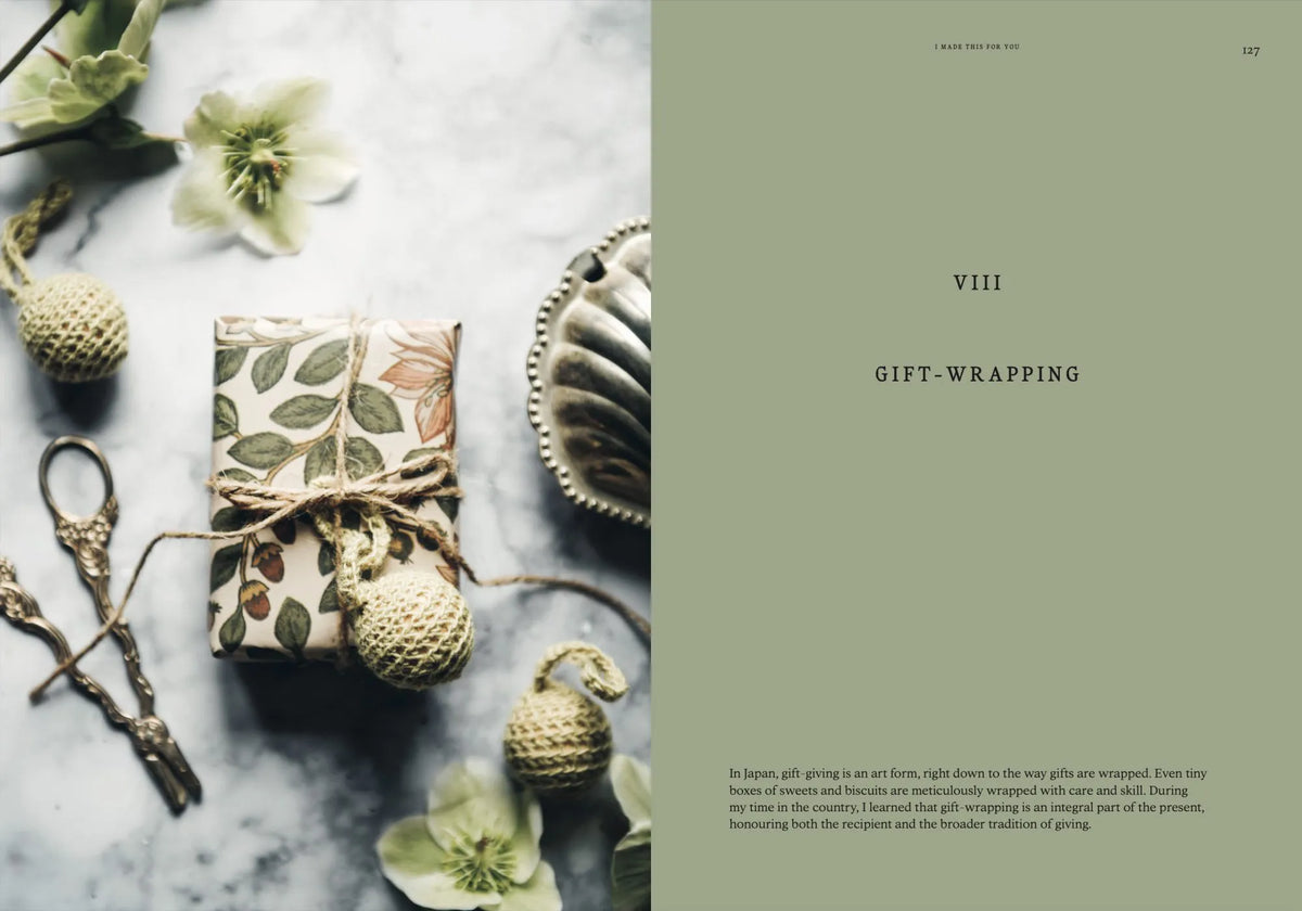 A copy of Cozy Publishing’s I Made This for You – Christmas Gift and Decorations Crafted with Love rests on a marble surface next to a floral-wrapped present with twine, scissors, and festive crafting decorations, perfectly capturing the charm of homemade Christmas gifts.