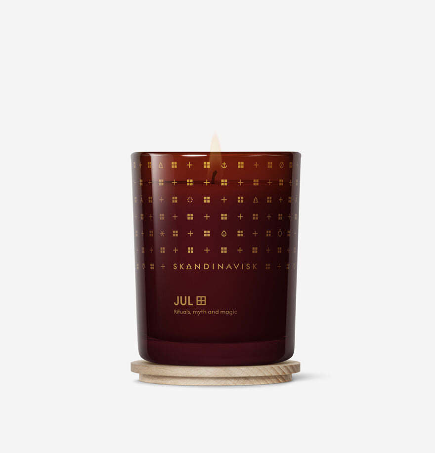 A lit Skandinavisk Seasonal Jul Scented Candle (50h, 200g) with a cozy gingerbread scent sits on a round wooden coaster against a white background, capturing the warmth of a Scandinavian Christmas.