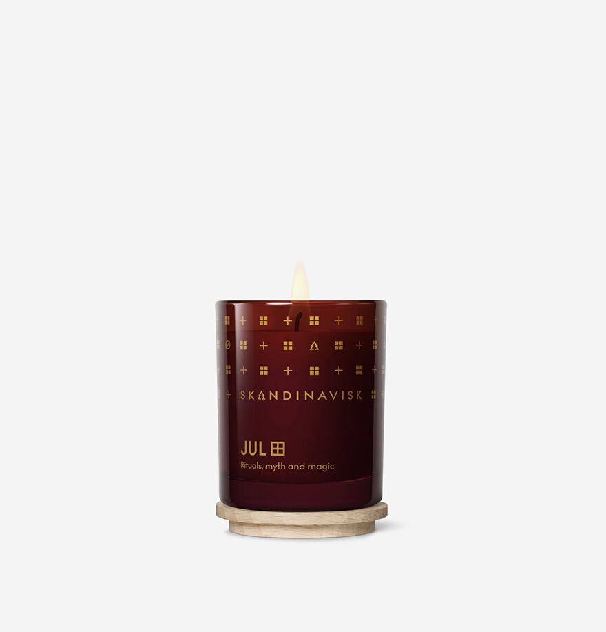 A lit Skandinavisk Seasonal Jul Scented Candle 65g in a dark red glass jar sits on a wooden coaster against a white background, filling the air with the cozy gingerbread aroma of Swedish rapeseed wax.