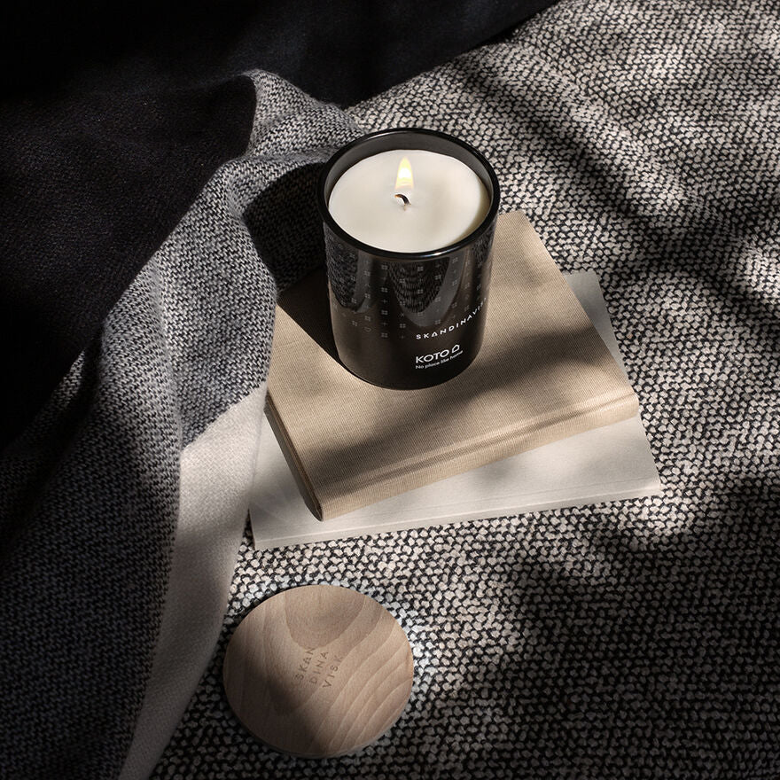 The Skandinavisk Scented Candle Koto 50h, 200g in a black glass container sits on two stacked closed books atop patterned fabric, with its wooden lid placed nearby.