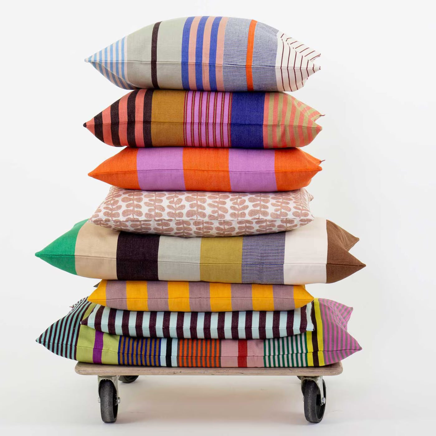 A stack of seven Olivia Striped handwoven cushion covers by A World of Craft (50 cm x 50 cm) in vibrant stripes and geometric patterns sits on a small wheeled cart against a plain white background.