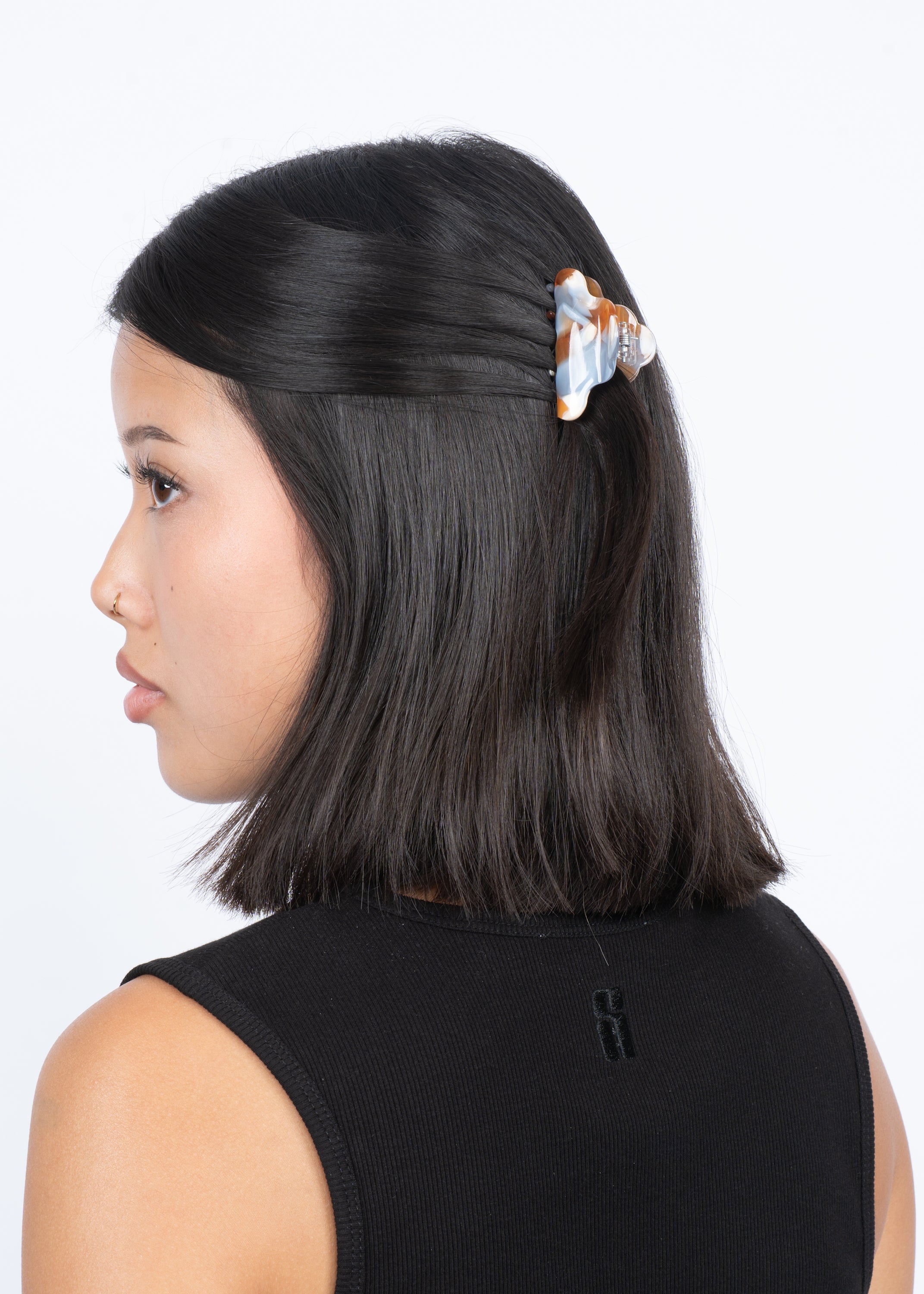 A woman with straight, shoulder-length black hair wears a black sleeveless top and has her hair partially clipped back with the Sui Ava Lene Icy Mini Hair Claw Clip in Salted Caramel. The background is white.
