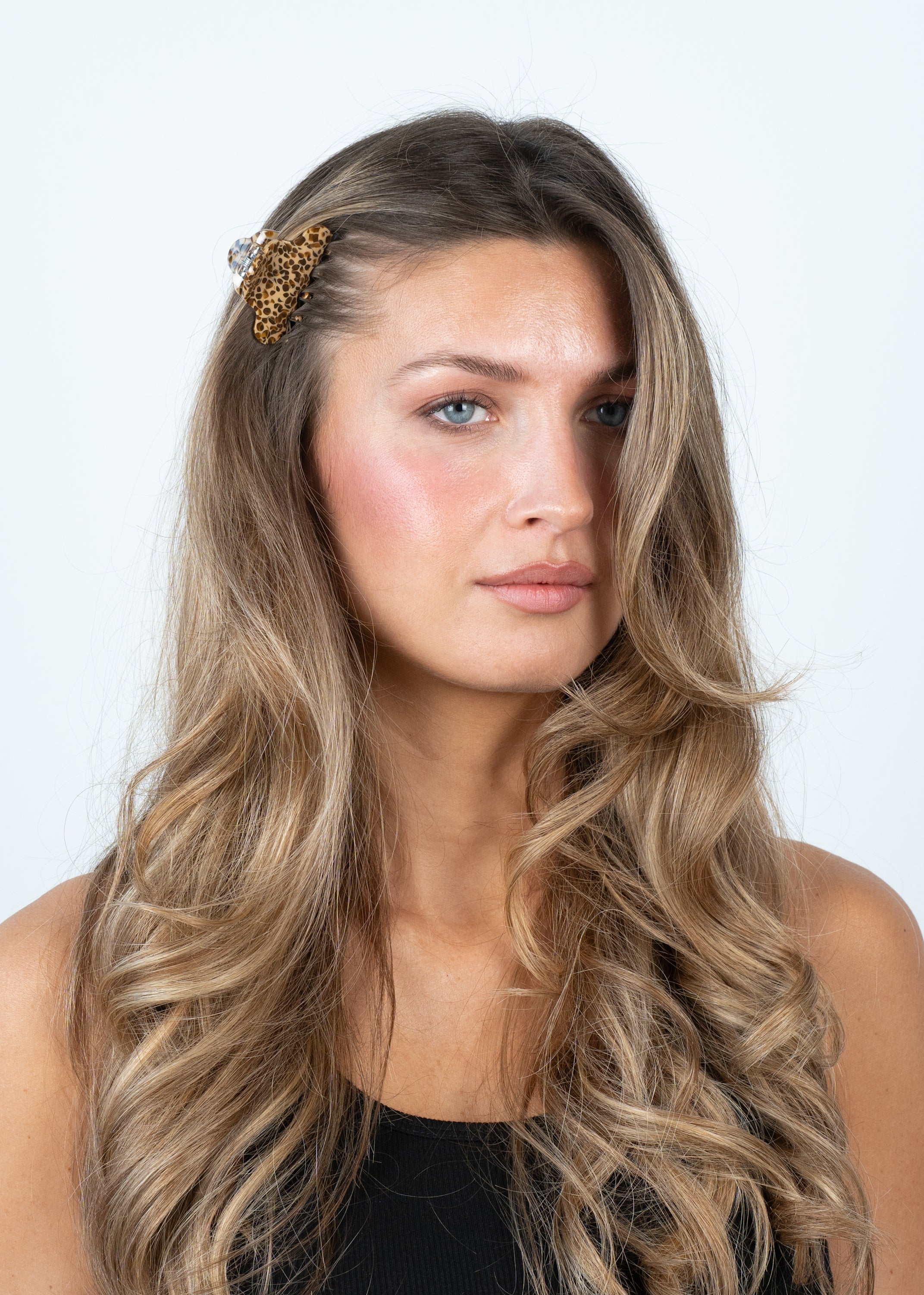 A woman with long, wavy blonde hair wears a black sleeveless top and the Sui Ava Lene Sable Mini Hair Claw Clip in Brown on the left side of her head. She looks slightly to the side against a plain white background.