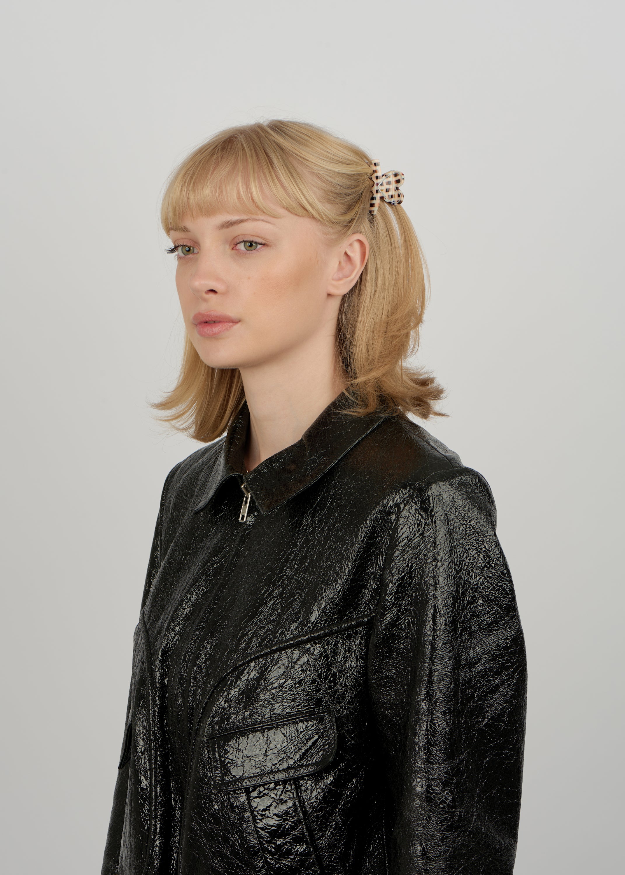 A young woman with blonde pigtails, wearing the Sui Ava Love on Repeat Mini Hair Claw Clip in Patchwork and a textured black jacket, stands against a plain light gray background.