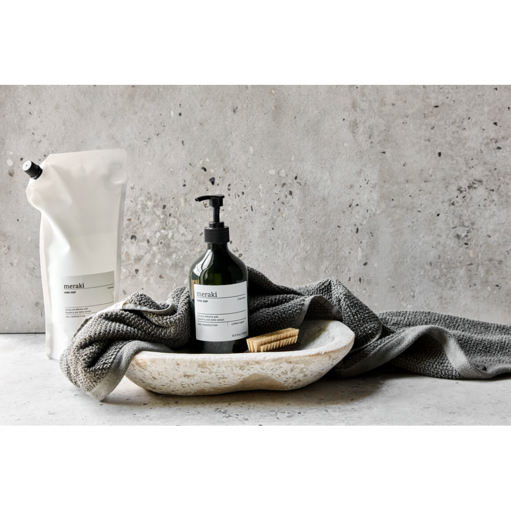 A stone tray holds a dark pump bottle of Meraki Hand Soap Organic, Linen Dew, a small wooden brush, and a gray textured towel. Behind the tray is a white refill pouch, all set against a gray stone backdrop for a minimalist, spa-like feel.