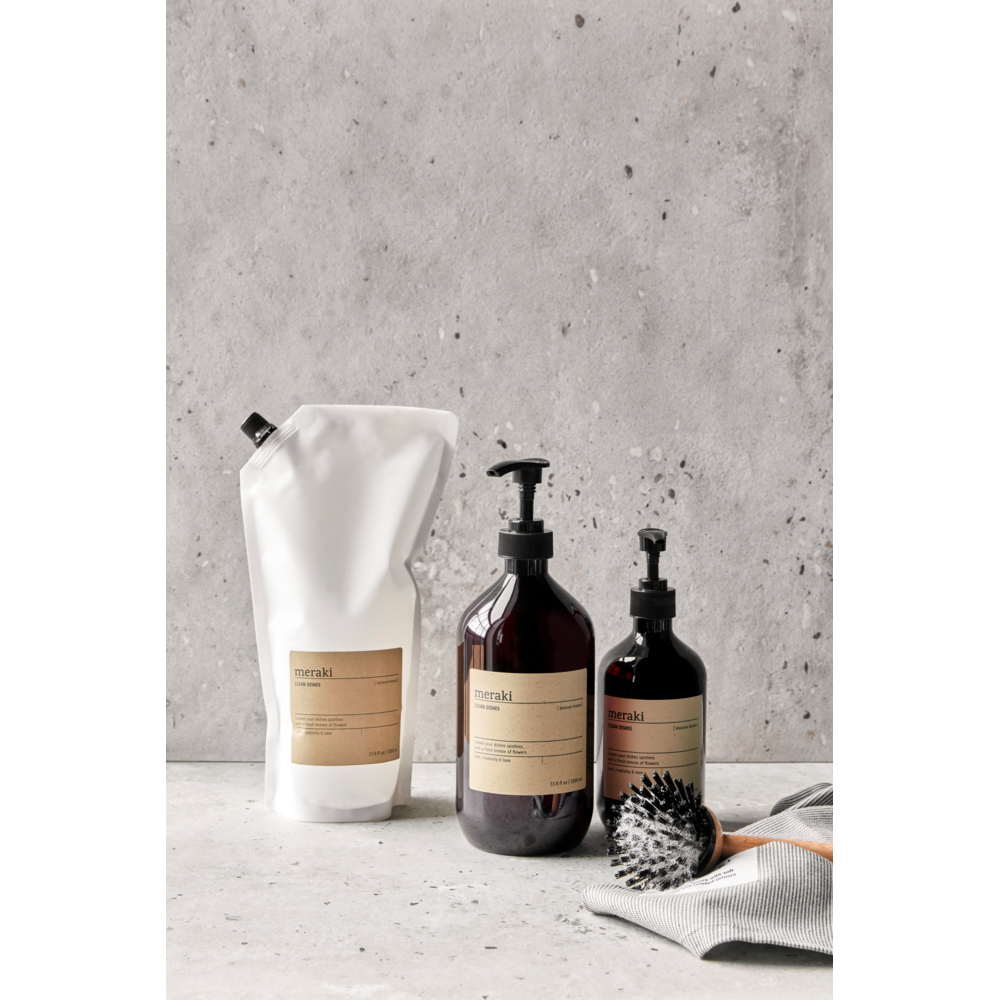Three Meraki Hand Soap Organic, Northern Dawn bottles—refill pouch, large pump, and small pump—are displayed on a gray surface with a brush and striped cloth in front of a concrete background.