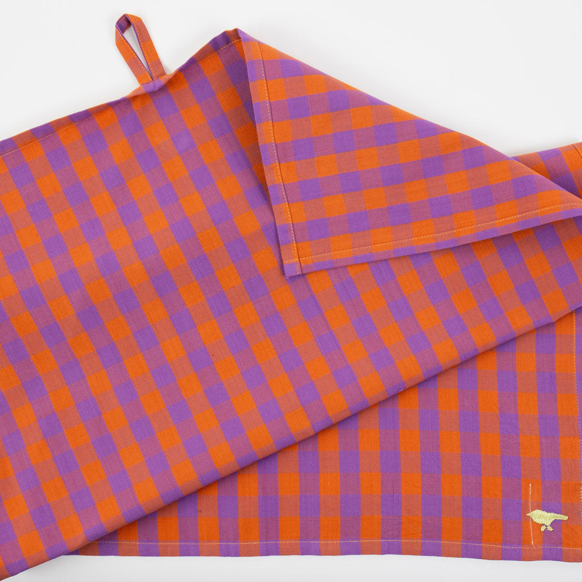 A folded Tea Towel, Nina Purple & Orange by A World of Craft, handwoven from organic cotton with an orange and purple checkered pattern, featuring a small loop on one corner and a subtle gold embroidered detail along the edge.