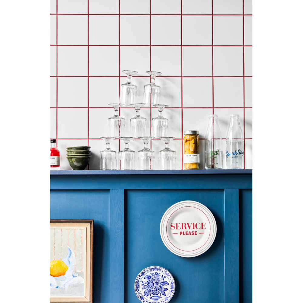 A row of upside-down glass cups stacked in a pyramid sits on a blue shelf against a white tile wall with red grout. Bottles, cans, and Nicolas Vahé Preserved Salted Lemons add decor alongside a SERVICE PLEASE plate.