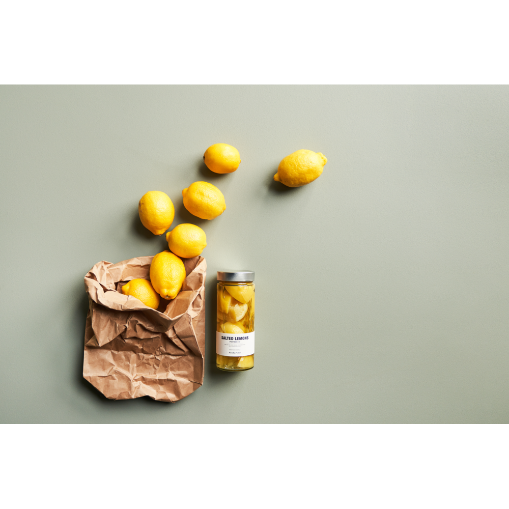 A crumpled brown paper bag with fresh lemons spilling out and a jar of Nicolas Vahé Salted Lemons, Preserved sits on a light green surface—ideal for adding bold Mediterranean flavor.