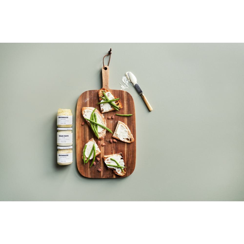 A wooden cutting board with sliced bread topped with cheese and green vegetables, a jar of Nicolas Vahé Mayonnaise with Black Truffle, two other seasoning jars, and a small cheese knife on a light grey surface.