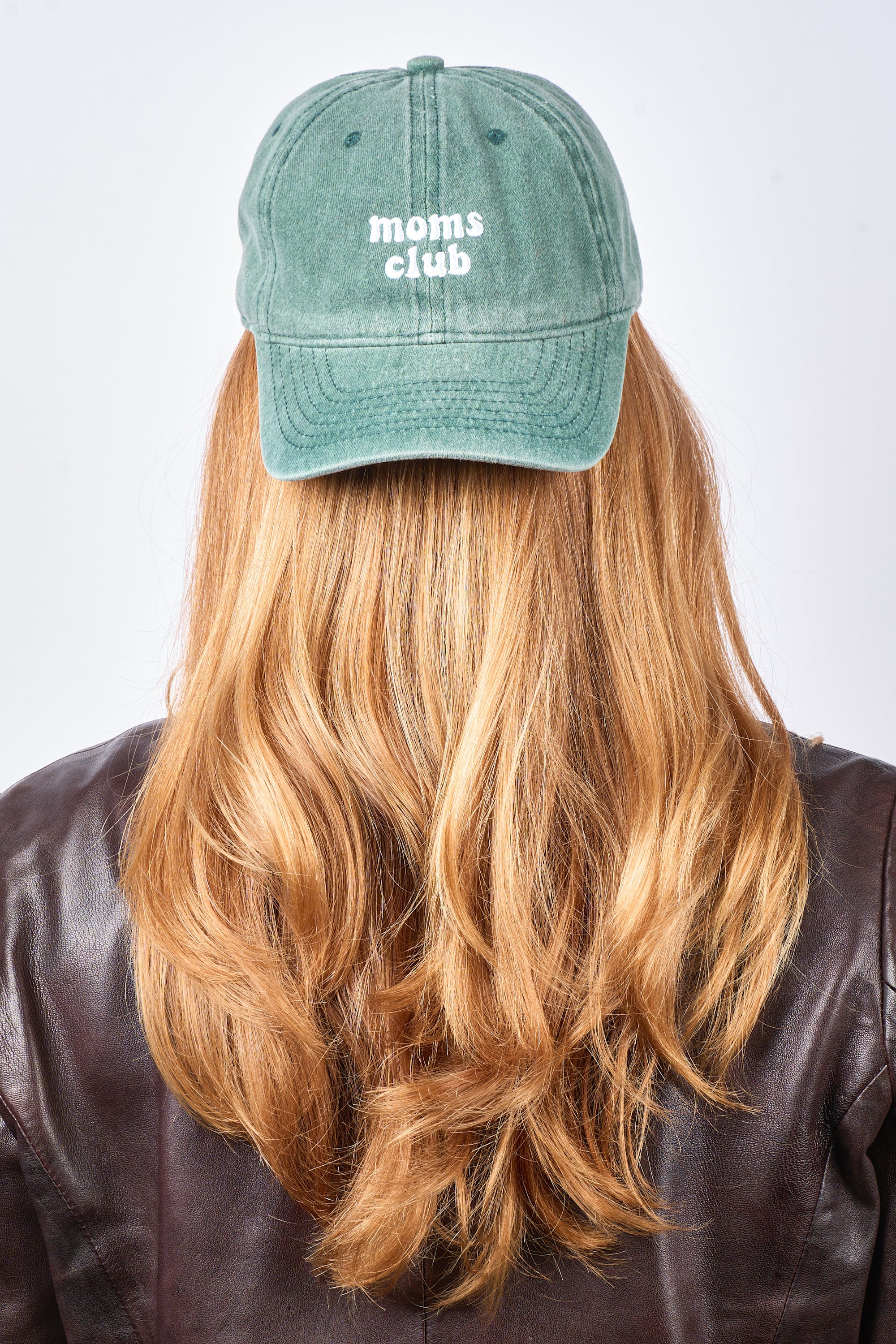 A person with long, wavy blonde hair is seen from behind wearing the Sui Ava Everyday Cap, Moms Club, in Green with “moms club” embroidered in white and paired with a brown leather jacket.