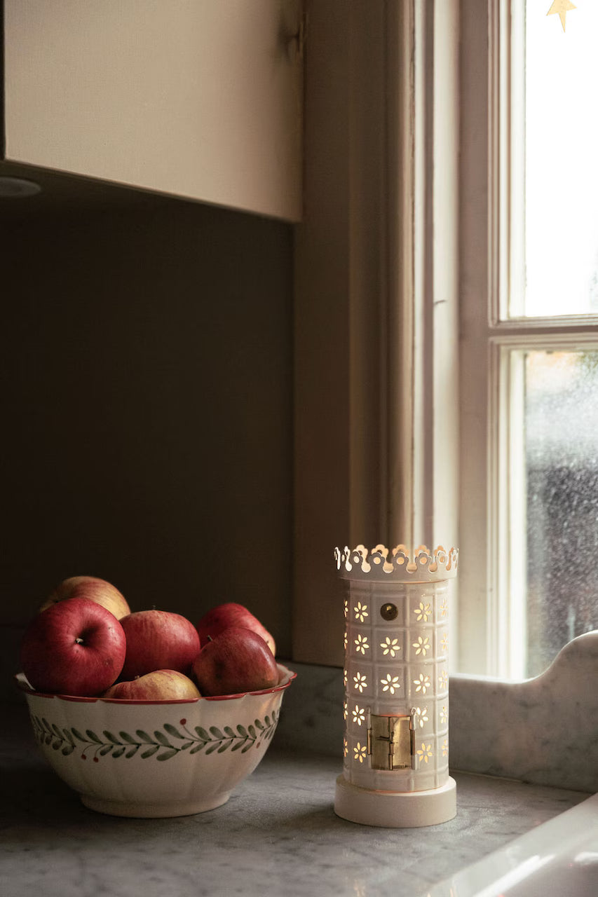 A bowl of red apples rests on the kitchen counter beside the Strömshaga Tea Light Holder, Tiled Stove in White, its soft glow reflecting off rainy windows and enhancing the cozy charm of the space.