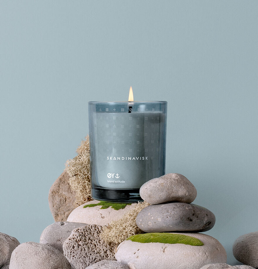 A lit Skandinavisk Scented Candle Øy, 200 g, in a glass holder sits on smooth gray stones and moss with a light blue background.