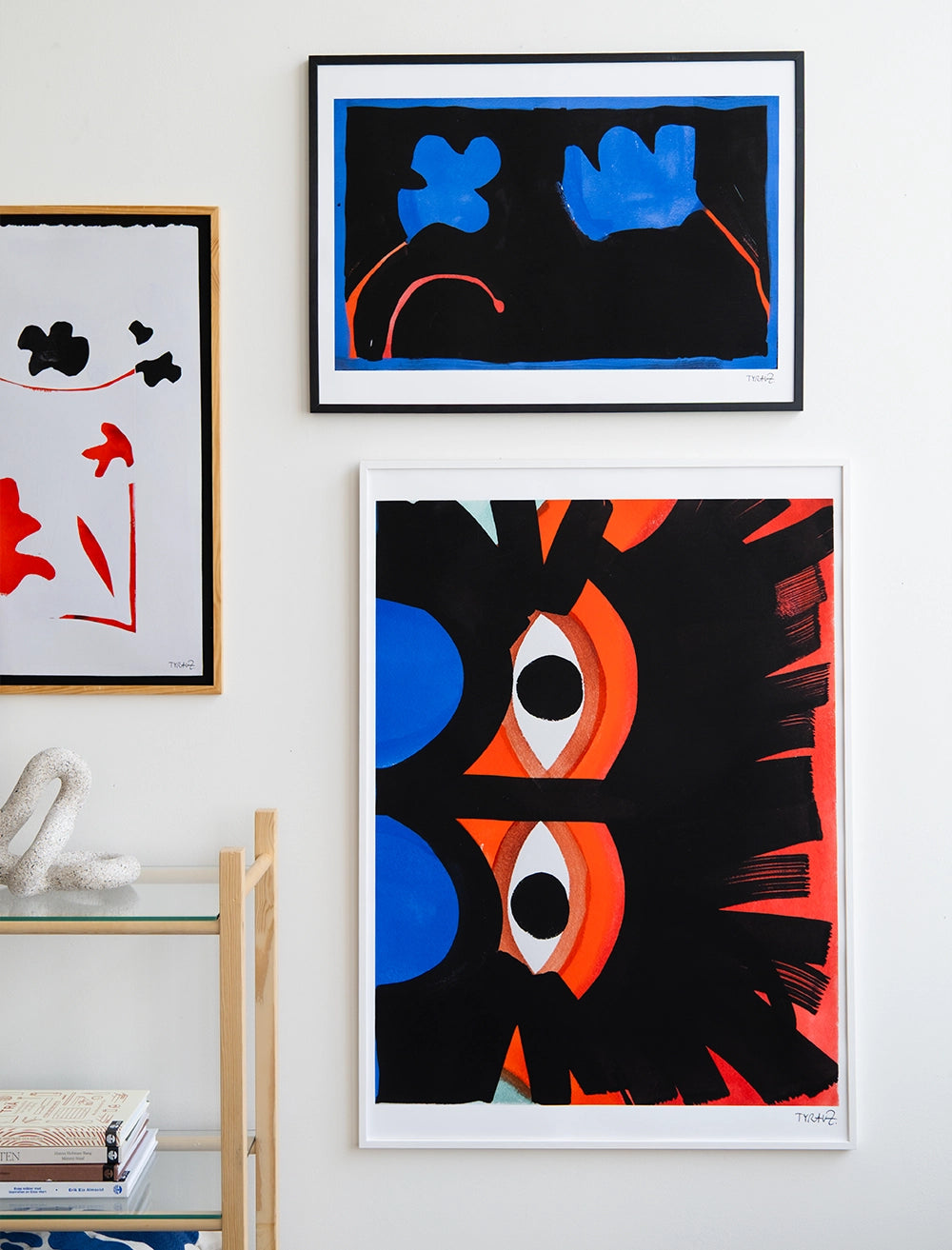 Eyes Print by Tyra von Zweigbergk (50x70 cm, Fine Little Day), featuring bold black, red, blue, and white abstract shapes, is displayed above a glass shelf with books and a decorative sculpture.