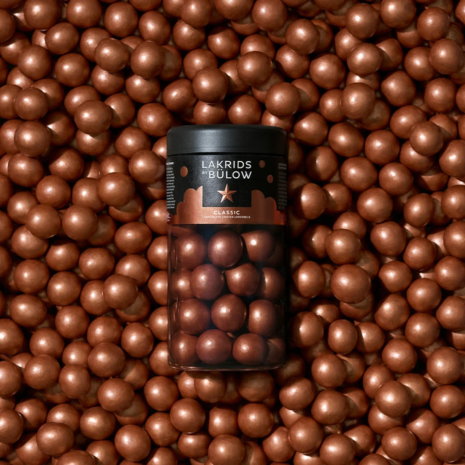 A jar of Lakrids by Bülow Classic Caramel Liquorice, Regular rests on a surface scattered with glossy caramel chocolate-coated liquorice balls, blending effortlessly with the treats around it—a true winter classic enhanced by the delicate crunch of sea salt flakes.