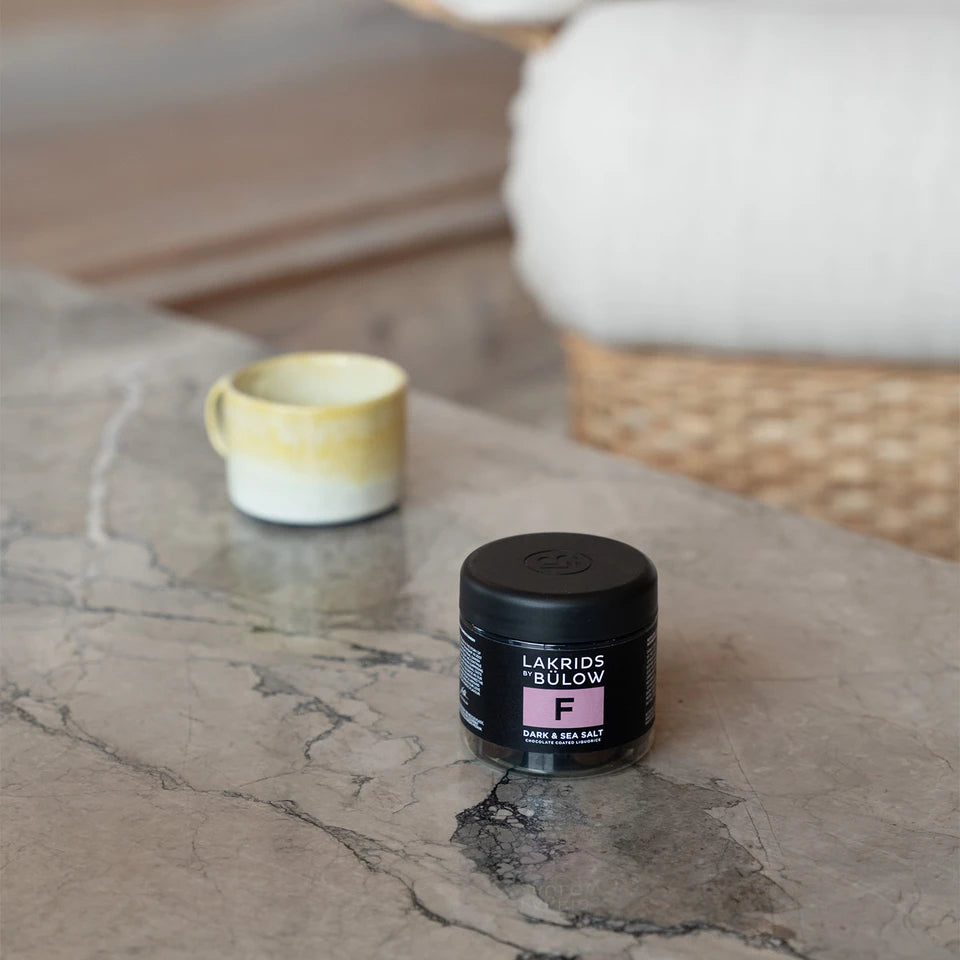 A small black jar labeled “Dark & Sea Salt Liquorice, Small, F” by Lakrids by Bülow sits on a marble table, its dark chocolate liquorice treats promising indulgence, with a yellow and white cup in the background near a wicker and white cushioned chair.