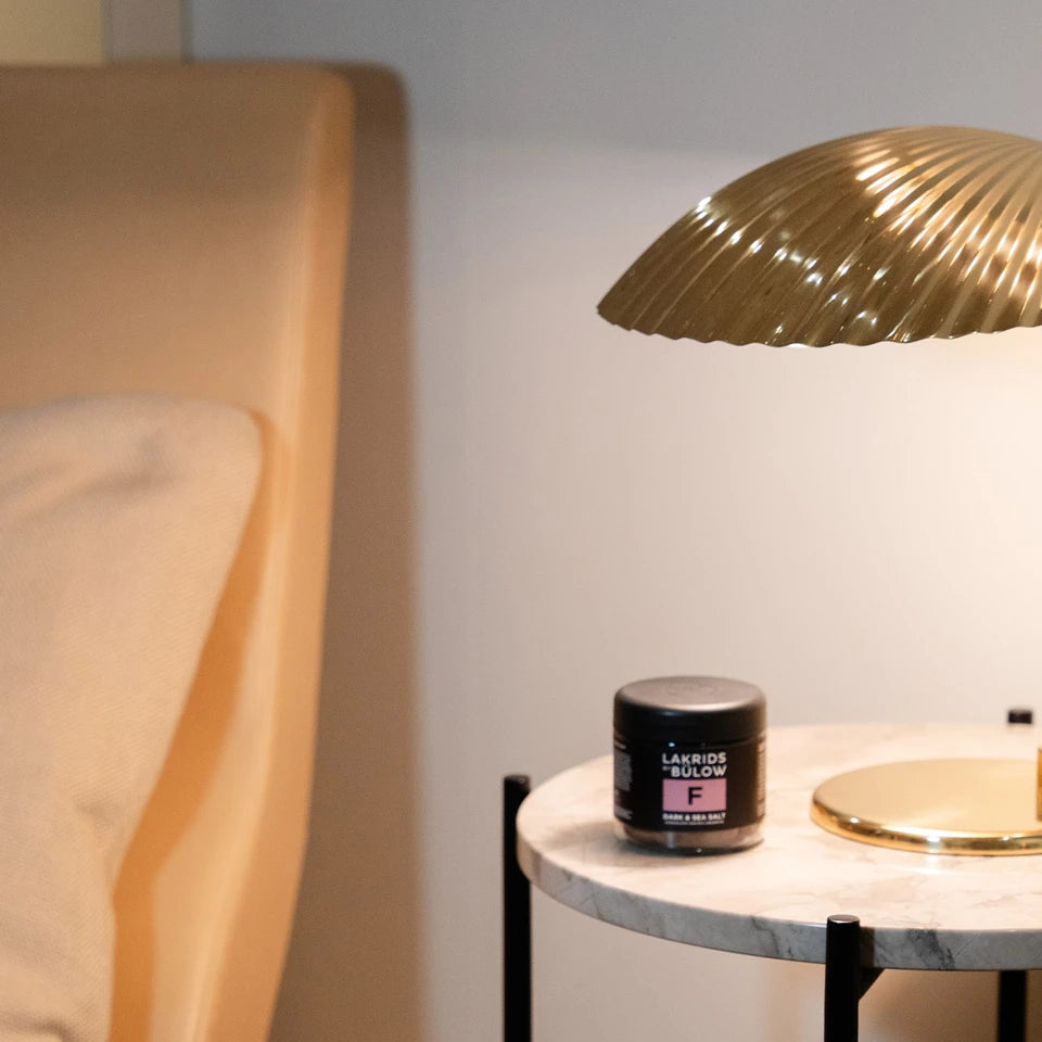 A modern gold table lamp and a small black jar of Lakrids by Bülow Dark & Sea Salt Liquorice, F, sit on a round marble side table next to a beige armchair with a cushion, set against a light-colored wall.
