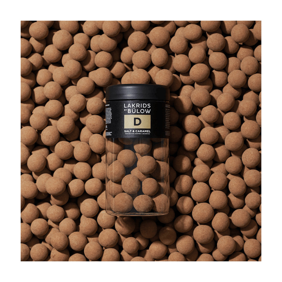 A jar of Lakrids by Bülow Salt & Caramel Liquorice, Regular, D is surrounded by numerous round, brown salt & caramel liquorice-coated candies, forming a textured and uniform backdrop.