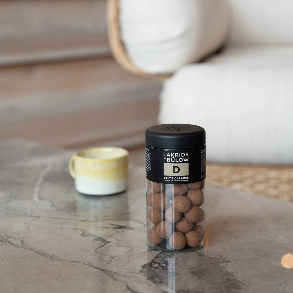 A jar of Lakrids by Bülow Salt & Caramel Liquorice, Regular, D rests on a marble table beside a yellow-green ceramic cup, with a sofa and wooden floor in the background.