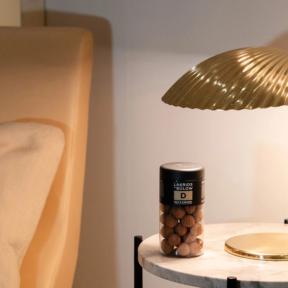 A gold lamp with a curved shade sits on a round marble table next to a bed. On the table is a jar of Lakrids by Bülow Salt & Caramel Liqourice, Regular, D, adding an indulgent touch of sweetness to the elegant setting.