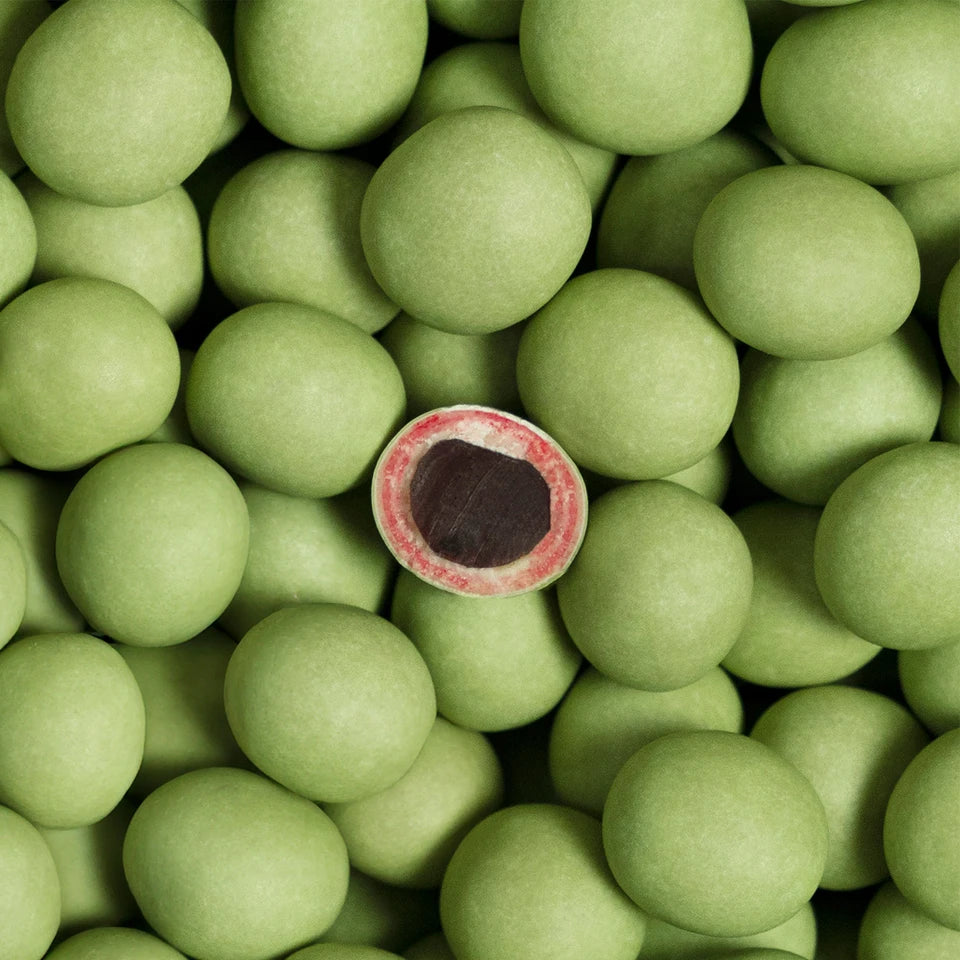 A group of Lakrids by Bülow Sour Strawberry Liqourice (Small, C) candies, featuring green candy-coated chocolates, with one cut open to display a dark chocolate interior encased in a layer of red liqourice beneath the green coating.