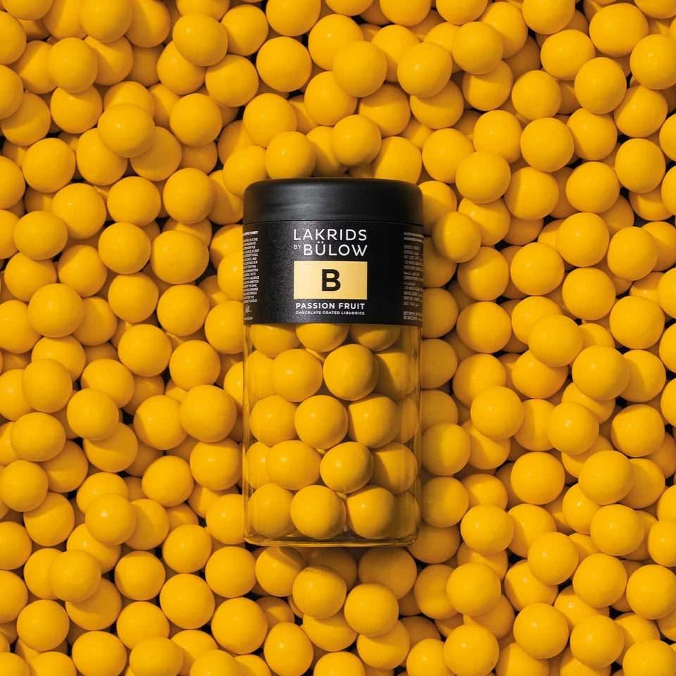 A jar of Lakrids by Bülow Passion Fruit Liqourice, Regular, B rests on a surface scattered with matching yellow spheres, creating a vibrant and cohesive yellow backdrop that highlights the sweet liqourice and passion fruit chocolate blend inside.