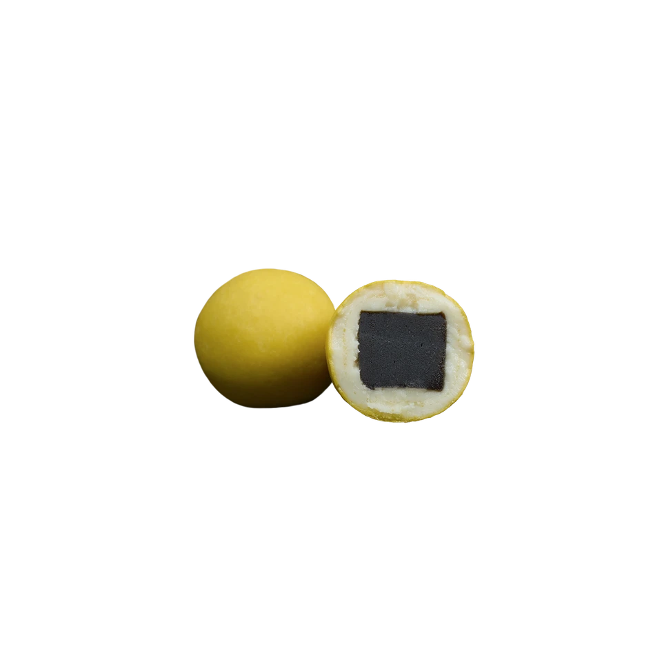 Two yellow spherical candies—one whole and one halved to reveal a white layer encasing a dark square center—are set against a plain white background. Lakrids by Bülow’s Passion Fruit Liquorice, Regular, B delights with its irresistible combination of sweet liquorice at the core and a luscious passion fruit chocolate coating, offering an intriguing blend of flavors and textures.