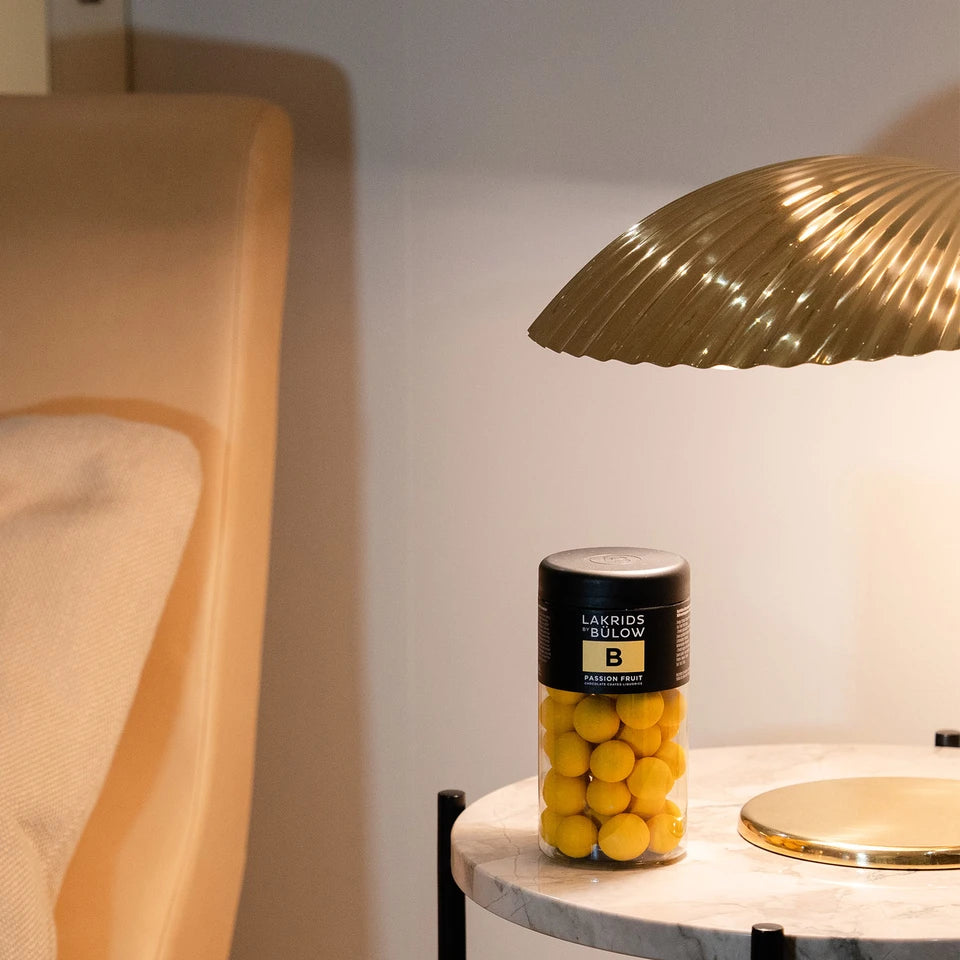 A jar of Lakrids by Bülow Passion Fruit Liquorice, Regular, B—filled with passion fruit chocolate and sweet liquorice—rests on a marble table beneath a gold lamp, beside a beige chair.