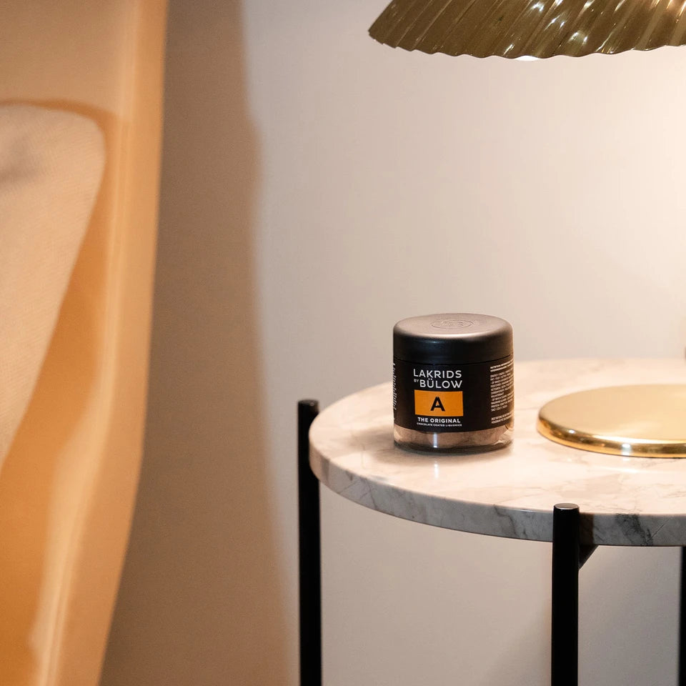 A jar labeled The Original Liquorice, Small, A by Lakrids by Bülow rests on a round white marble side table with black legs, positioned next to a gold lamp. In the background, a beige chair and softly lit wall create an inviting setting that echoes the indulgent blend of milk chocolate and sweet liquorice in every bite.