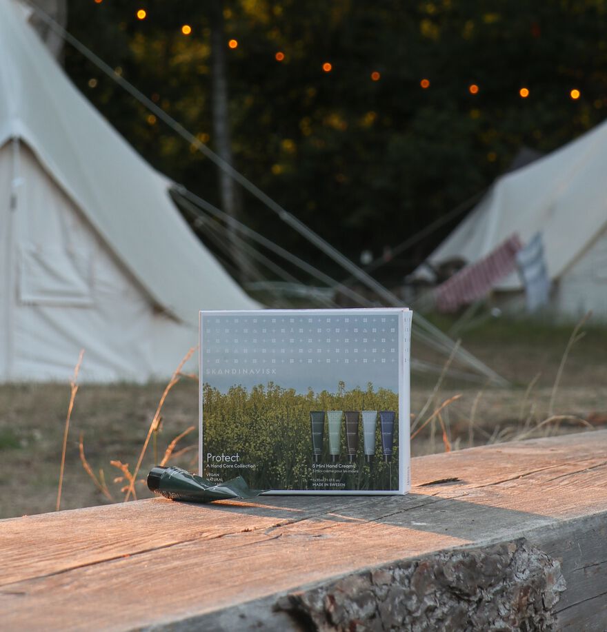 A set of Skandinavisk Giftset Mini Hand Cream Protect, featuring five tubes inspired by Skog, Fjord, Oy, Hav, and Hygge, rests on a wooden surface outdoors. White tents and string lights in the background create a rustic campsite scene, while the packaging emphasizes natural Scandinavian ingredients like organic aloe vera to enhance the fresh and wholesome atmosphere.