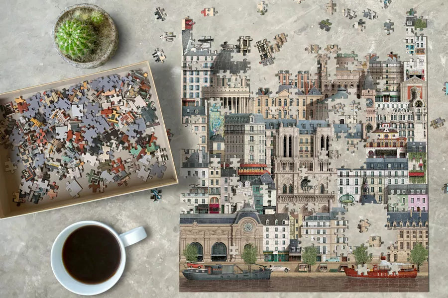 A partially completed Martin Schwartz Paris Puzzle (1000 pieces) lies on a table, with loose pieces in a box to the left, and a cup of black coffee plus a small potted cactus nearby.
