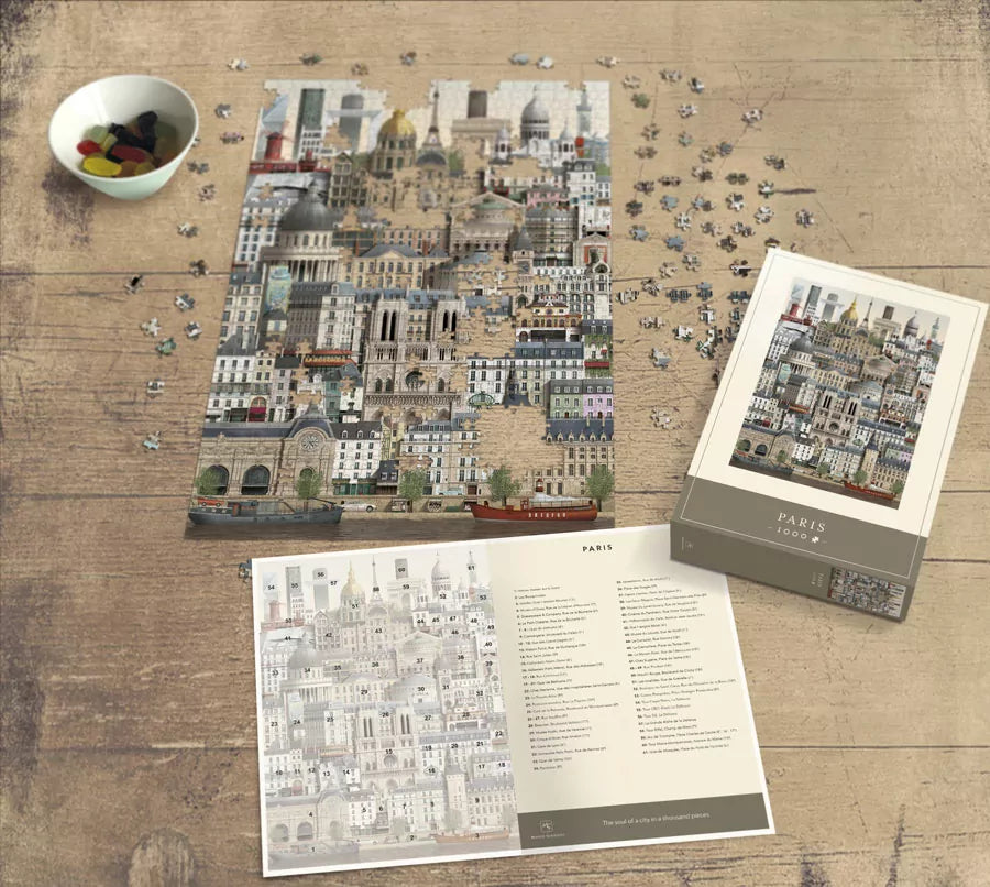 A partially completed Martin Schwartz Paris Puzzle (1000 pieces) rests on a wooden table with loose pieces, a bowl of snacks, the puzzle box displaying the finished image, and an open booklet with text and illustrations nearby.